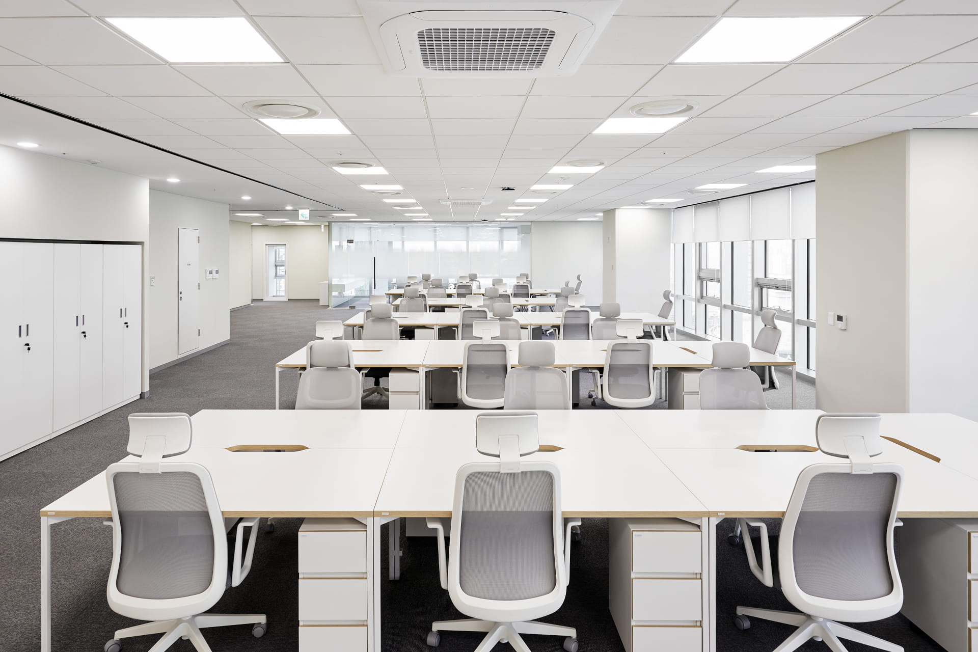 화이트 책상과 메쉬 소재 사무용 의자가 일렬로 배치된 넓고 쾌적한 오픈 오피스 사무 구역.jpg