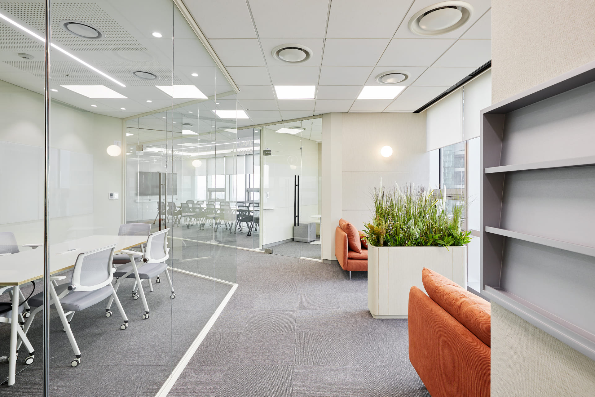 투명한 유리 가벽으로 개방감을 준 회의실과 오렌지색 소파가 배치된 쾌적한 오피스 통로 전경.jpg