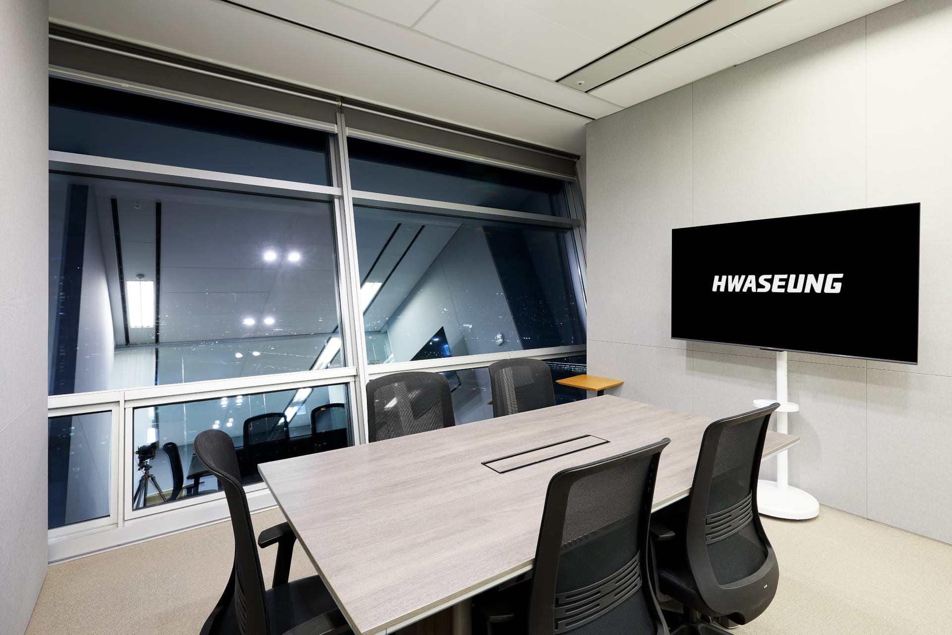 야경이 보이는 대형 창문 옆에 위치한 4인용 목재 미팅 테이블과 화승 로고가 송출되는 벽걸이 TV.jpg