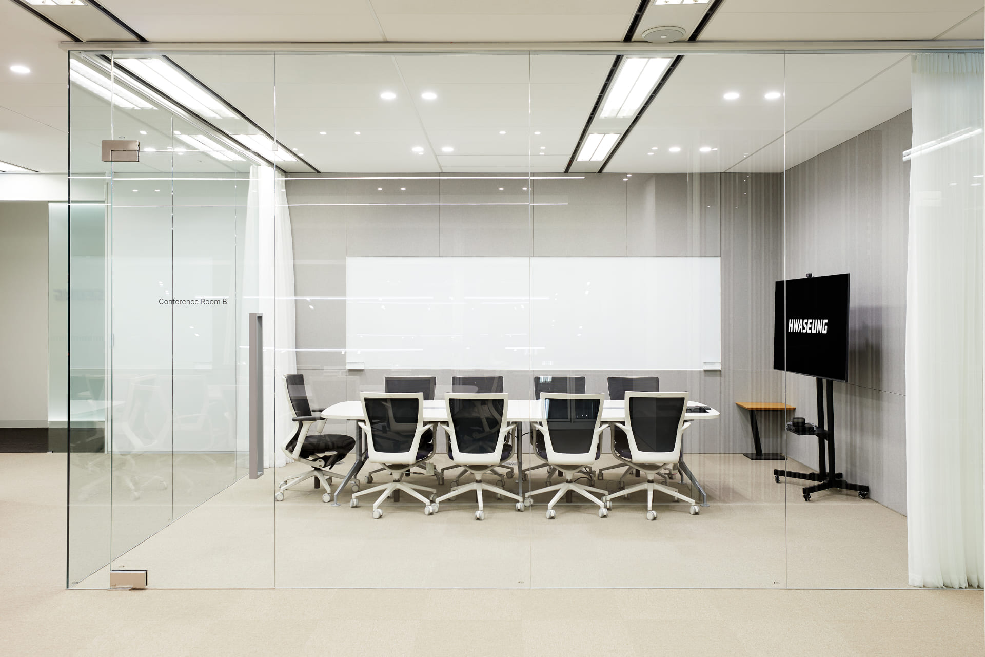 투명한 유리 벽체로 개방감을 준 컨퍼런스룸 B 내부의 화이트 테이블, 블랙 사무용 의자, 대형 모니터 구성.jpg