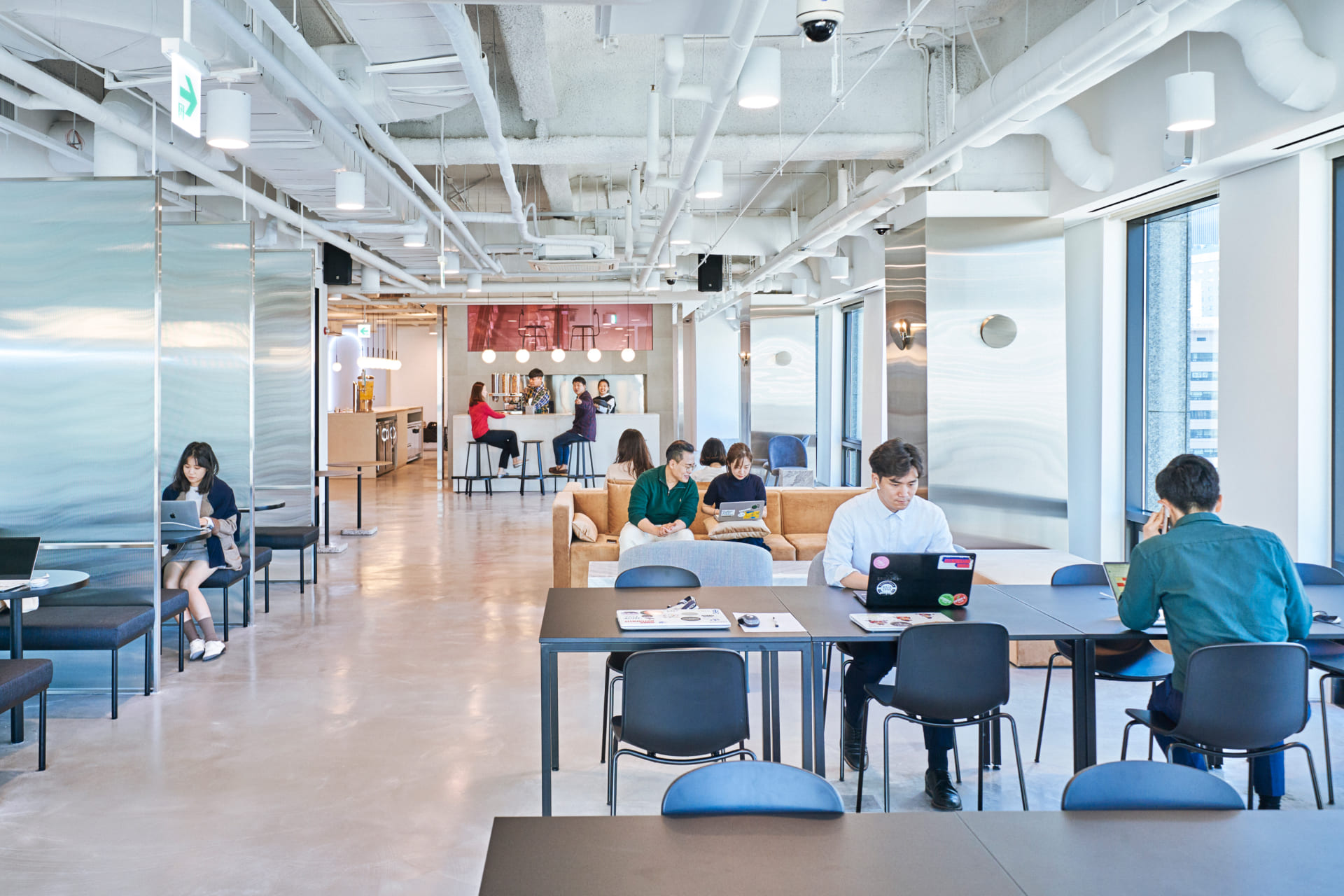 개방형 천장 아래에서 자유롭게 노트북으로 업무를 보거나 대화하는 이용자들로 활기찬 공유오피스 메인 홀 전경.jpg