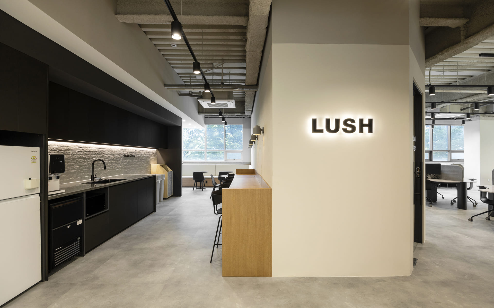 LUSH 로고가 빛나는 미니멀한 오피스 카페테리아로 검은색 주방 가구와 우드 바 테이블이 조화를 이룬 공간.jpg
