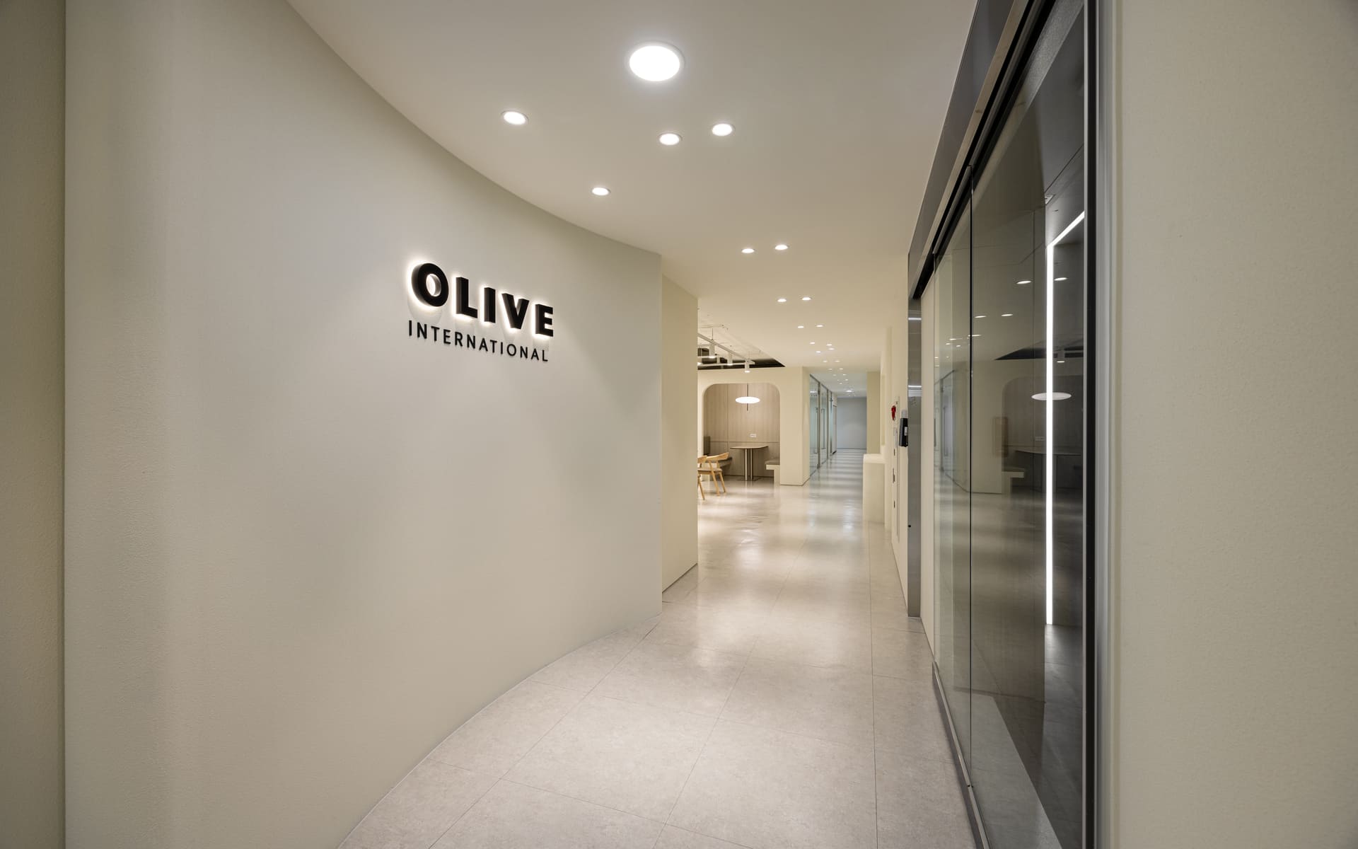 OLIVE INTERNATIONAL 로고가 부착된 부드러운 곡선 벽면과 은은한 매립등이 설치된 사무실 진입 복도.jpg