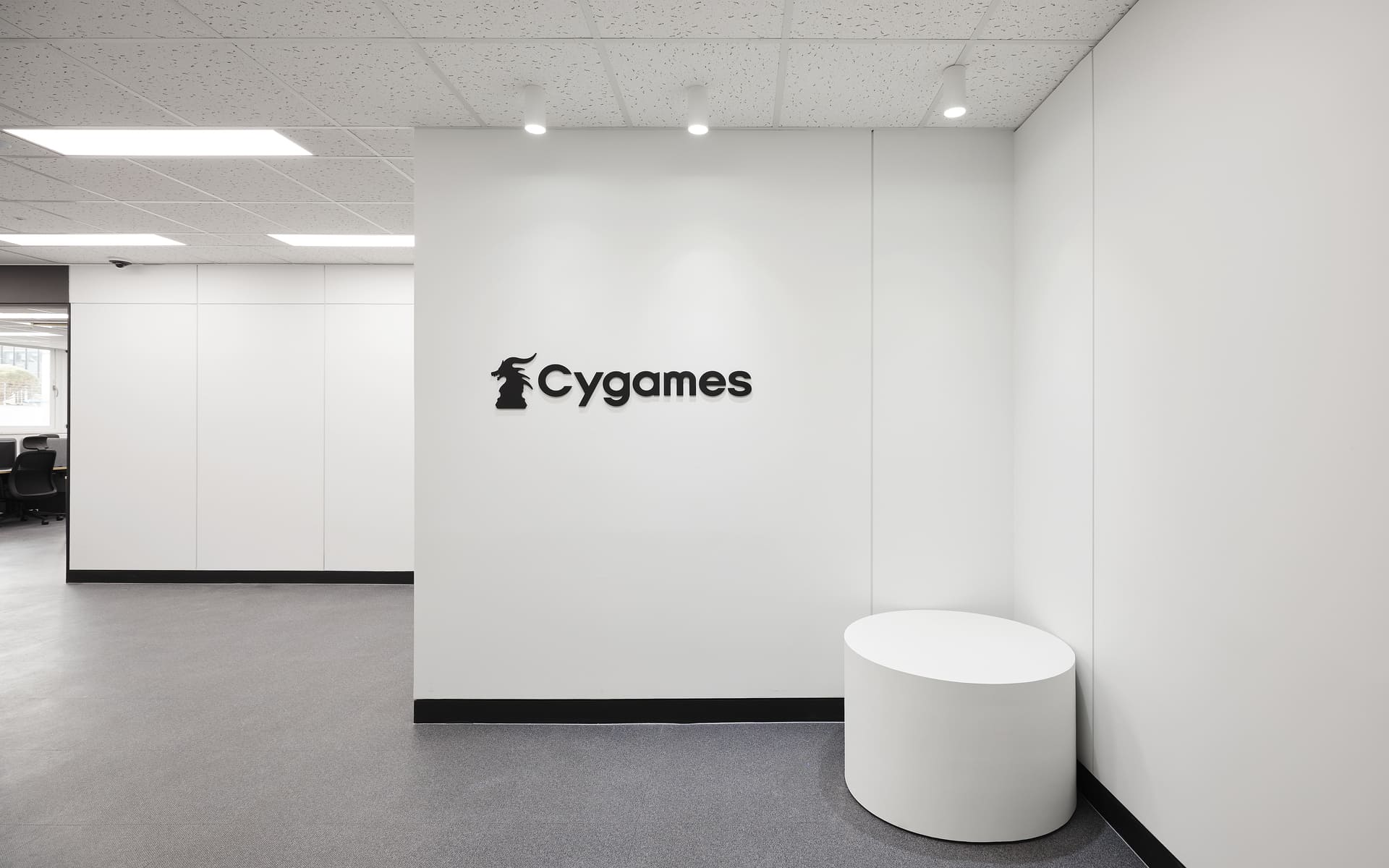 화이트 톤의 미니멀한 복도 벽면에 부착된 Cygames 로고와 심플한 화이트 원형 스툴 인테리어.jpg