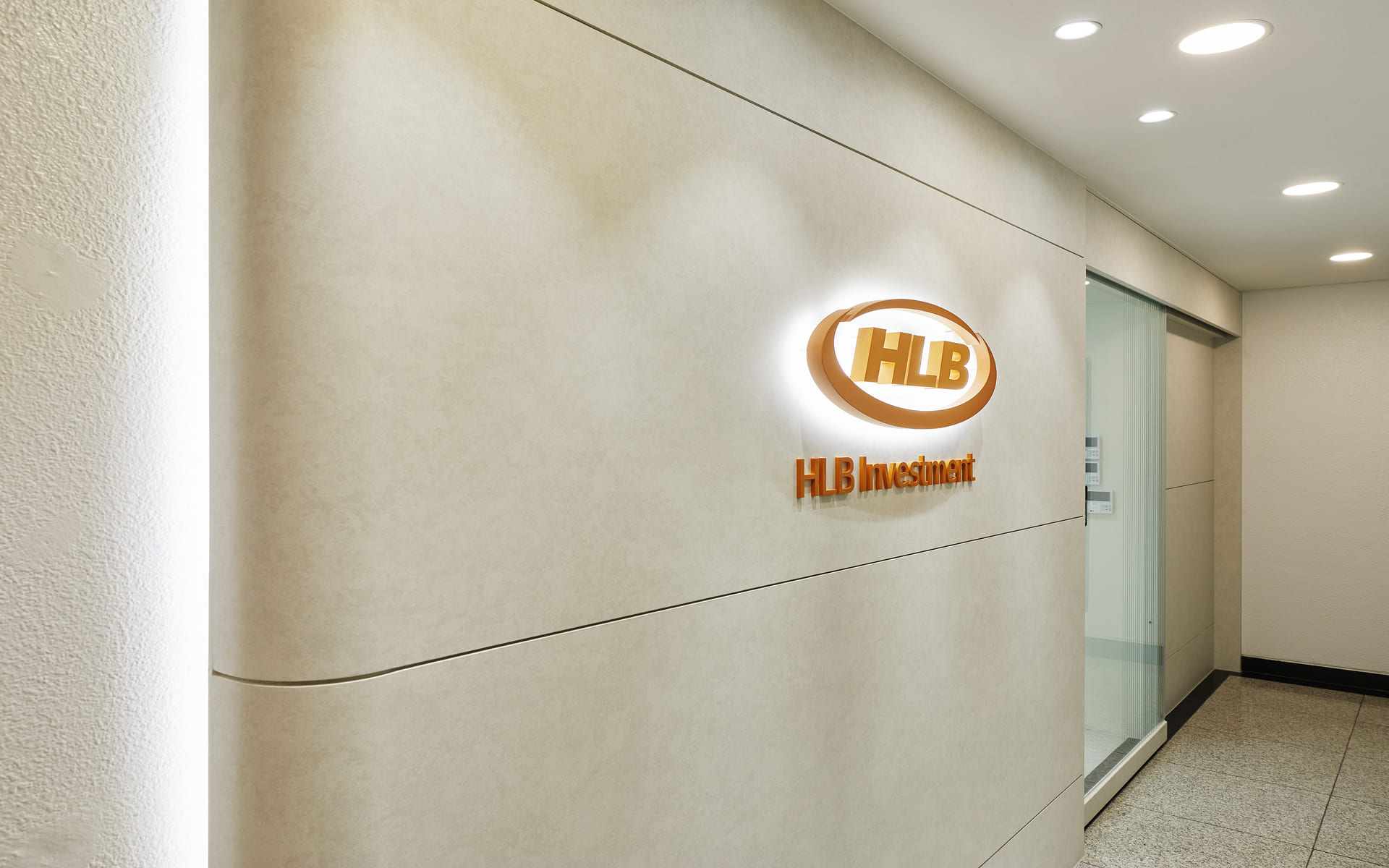 은은한 간접 조명과 곡선형 벽면에 HLB Investment 로고가 부착된 세련된 기업 입구 인포메이션.jpg