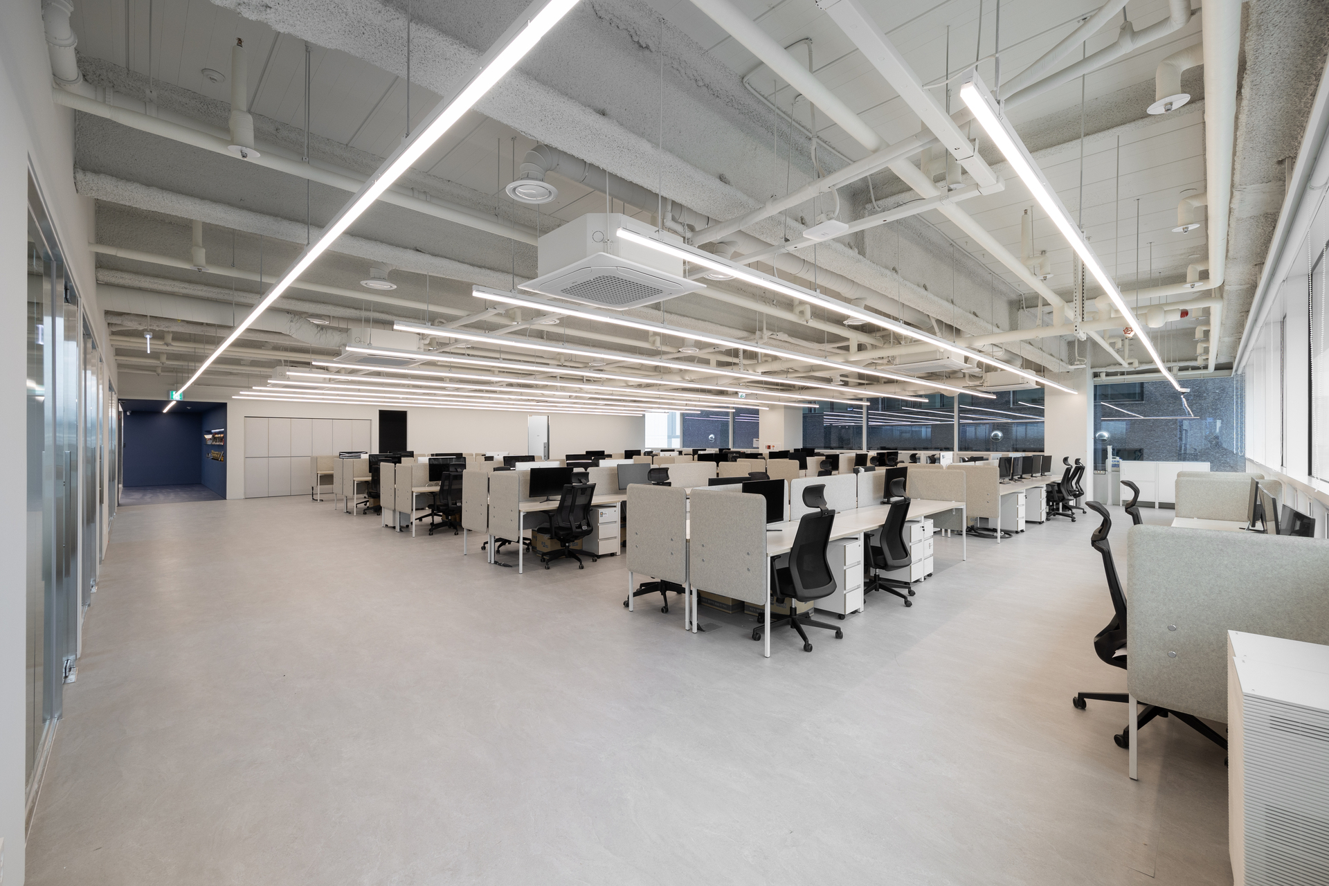 노출 천장과 화이트 조명 라인이 돋보이는 탁 트인 대규모 오피스의 개방형 워크스테이션 사무 공간 전경.jpg