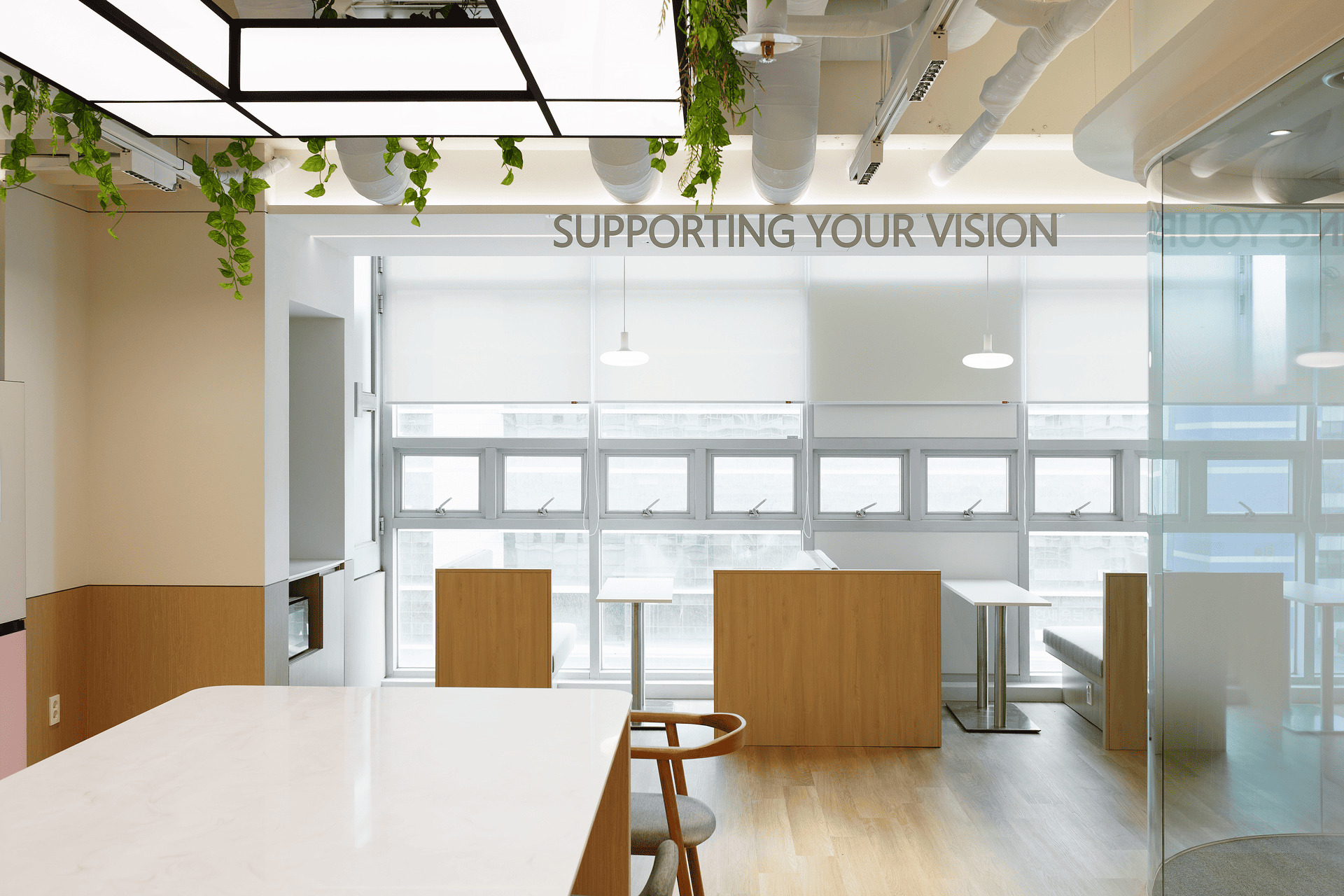 창가 밖으로 SUPPORTING YOUR VISION 문구가 보이는 통유리창 미팅 부스와 아일랜드 식탁이 들어선 라운지.jpg